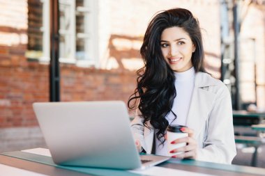 Koyu dalgalı saçlı bir kenara kafede temiz hava açık havada hayran paket servisi olan restoran kahve içme sınıfları sonra dinlenme ararken ifade memnun onu laptop haber okuma gülümseyen kız öğrenci
