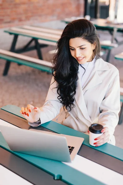 Mensen en levensstijl concept. Bovenaanzicht van schattige brunette vrouw in witte jas met rode manicure zittend aan tafel werken met laptopcomputer, gelukkig op zoek naar scherm heerlijke koffie drinken — Stockfoto