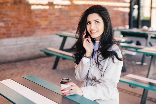 Linda mulher de cabelos escuros com pele pura, olhos brilhantes escuros e lábios cheios segurando café takeaway e smartphone conversando com seu namorado ter olhar satisfeito. Jovem empreendedora descansando — Fotografia de Stock