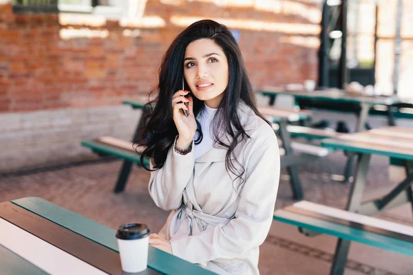 Portret van mooie vrouw met een aantrekkelijk uiterlijk, het dragen van witte formele kleding spreken over mobiele telefoon en drinken afhaalmaaltijden koffie opzoeken met doordachte expressie. Gezichtsuitdrukkingen — Stockfoto