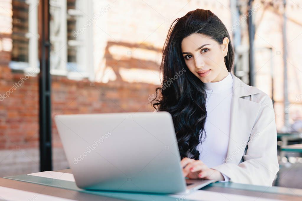 Portrait of beautiful brunette female freelancer typing a message via email using laptop while sitting on terrace looking at camera with confident look. Young lady working with computer outdoors