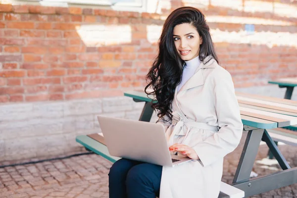 Люди, технологии, коммуникационная концепция. Красивая женщина с темными волнистыми волосами в белом плаще сидит на скамейке над кирпичной стеной держа ноутбук общаться онлайн с помощью бесплатного интернета — стоковое фото