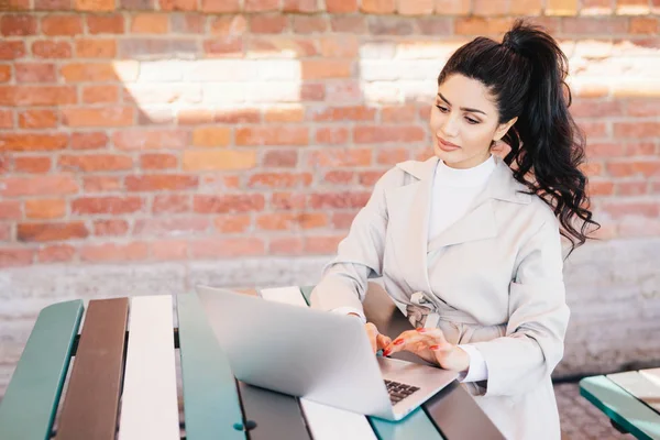 İnsan ve teknoloji. Dizüstü bilgisayar, manifatura sokulgan yolu ile sosyal ağlar tuğla duvarın üzerinden rahat kafede masada oturan yazarak Beyaz önlük giymiş at kuyruğu siyah saçlı genç güzel kadın bağladılar