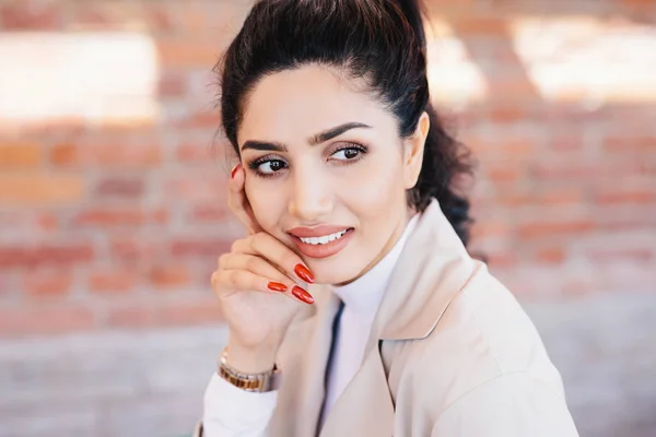 Close up of glamour brunette female having thin eyebrows, dark eyes with long eyelashes, pure skin, full well-shaped lips, red manicure wearing elegant watch on hand and white jacket looking aside — Stock Photo, Image