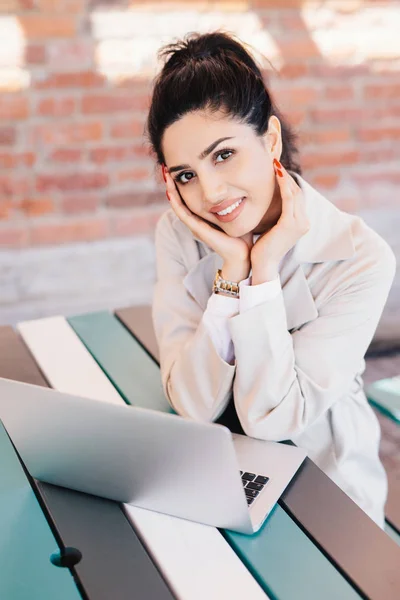 Retrato interno de uma jovem blogueira caucasiana bem-sucedida escrevendo um novo post para seu blog popular usando notebook enquanto se senta na mesa sobre o fundo da parede de tijolo. Sorrindo senhora bonita com unhas vermelhas — Fotografia de Stock