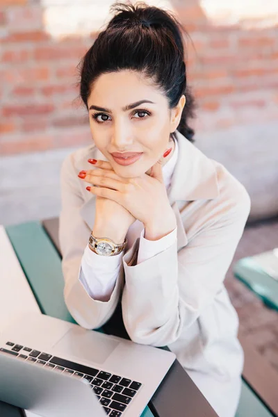 Young brunette businesswoman with charming eyes, gentle hands with red manicure wearing watch on hand and white coat holding hands under chin sitting near her laptop having rest after hard work