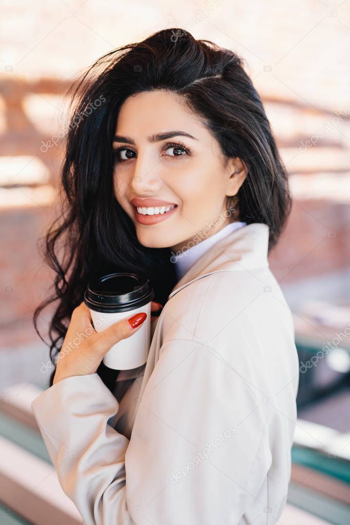 Good-looking woman with luminous dark hair, warm eyes and well-shaped lips drinking hot tea from paper cup while standing outdoors. Woman having rest enjoying drinking coffee. Rest and lifestyle