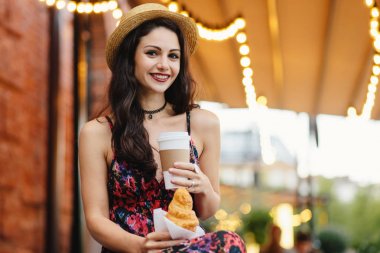 İnsanlar, yemek, dinlenme ve yaşam tarzı kavramı. Uzun saçlı esmer kadın yazlık elbise ve şapka, paket servisi olan restoran kahve ve eatting kruvasan açık kafede otururken içme giyiyor rahat dinlenme