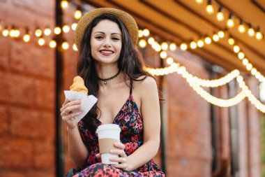 Genç kadın modeli ile koyu renk gözleri, kırmızı yazlık elbise ve eller paket servisi olan restoran kahve ve kruvasan, açık restoranda kahvaltı holding şapkası giyen dudakları boyalı. Gıda ve gevşeme kavramı