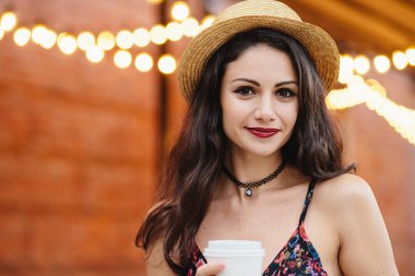 Şirin esmer erkek saf sağlıklı deri, koyu renk gözleri, kırmızı dudaklar ile hasır şapka ve paket servisi olan restoran kahve terasında poz tutan elbise giyiyor. Genç erkek kahve molası verdikten sonra zor işi ofiste