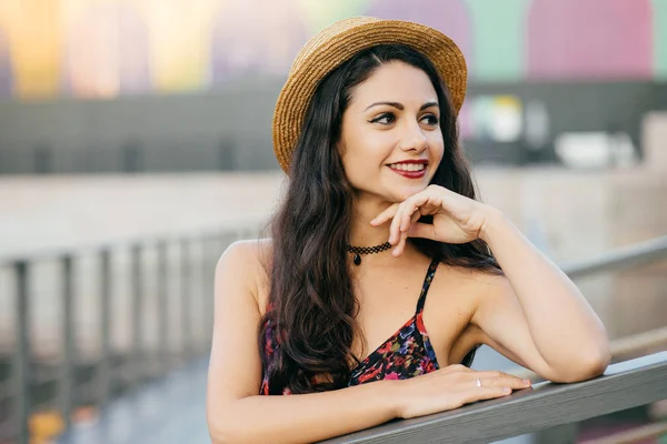 Young female model with long dark thick hair standing at bridge having dreamy expression looking into distance, wearing summer hat and dress resting outdoors. People, relaxation, beauty concept — Stock Photo, Image