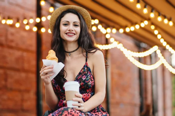 Genç kadın modeli ile koyu renk gözleri, kırmızı yazlık elbise ve eller paket servisi olan restoran kahve ve kruvasan, açık restoranda kahvaltı holding şapkası giyen dudakları boyalı. Gıda ve gevşeme kavramı