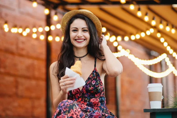 Şapka, elbise ve kolye, lezzetli kruvasan neşeli göz olan holding giyen görünüm, çekici güzel kadın siyah saçlı. Mutlu genç güzel kadın terasında Kahvaltı