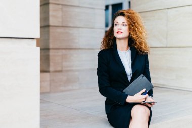 Gür saçlı, mavi gözlü, kırmızı dudaklar, yakışıklı genç iş kadını tutan eller cep kitabı ve modern tablet bilgisayar, siyah takım elbise giyen bir kenara düşünceli ifade ile seyir