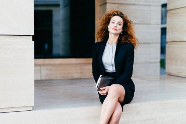 Çok güzel kadın modeli gür dalgalı saçlı, kırmızı dudaklar ve parlayan gözleri, resmen tablet cep kitabı, güvenle kameraya bakarak ile tutarak bacaklarını geçerken giyinmiş. İş kavramı