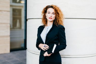 Resmi elbiseler giymiş genç güzel giyimli kadın resmen, cüzdan ile kalem tutan, gür saç modeli sahip, office builduing duran. Çalışma başarılı oldukça kadın ofis çalışanı