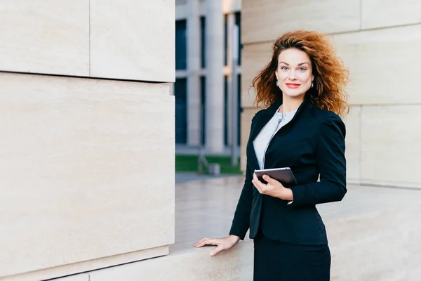 Cute female freelancer with luxurious light hair, blue eyes, red rouged lips and pure skin, wearing black formal suit, holding electronic gadget n hands, going on meeting with her business partners — Stock Photo, Image
