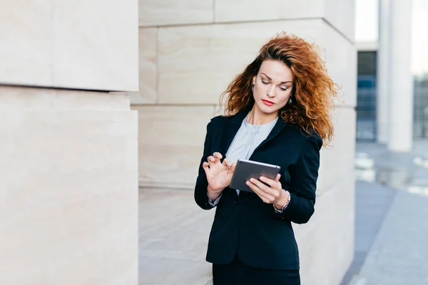 Elegant lady with curly hair, wearing black suit, typing messages or making business report while using tablet computer. Female entrepreneur working on new business project. Business and career