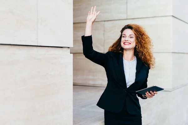 Smiling happy businesswoman dressed formally, holding in hands modern tablet computer, waving with her hand while noticing her companion, being glad to see him. Woman with curly hair saying `hello` — Stock Photo, Image