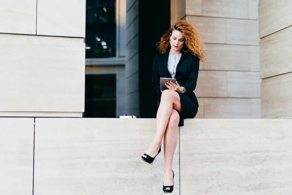 Young female model with curly hair, wearing elegant suit and high-heeled shoes, having slender legs, using modern tablet for communication. Young freelancer working outside, looking seriously — Stock Photo, Image