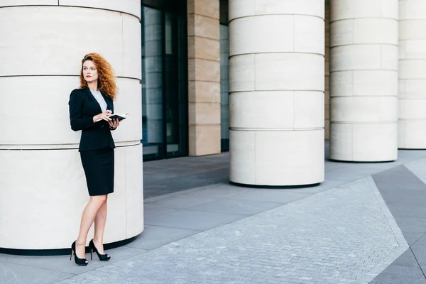 Horizontal portrait of good-looking businesswoman dressed in formal clothes and black shoes with high heels, holding pocket book, looking aside with thoughtful expression. Career and business concept — Stock Photo, Image