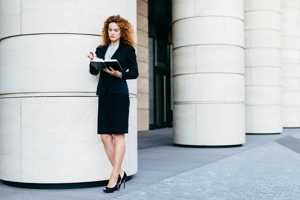 Slim young businesswoman in elegant clothes and high-heeled shoes, having concentrated look in her notebook, finding spare date for meeting. Serious female freelancer with her diary book and pen — Stock Photo, Image