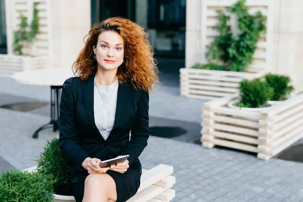 Beautiful gorgeous woman with wavy bushy hair, wearing formal suit, sitting crossed legs at outdoor cafe, holding tablet computer while waiting for someone, looking at camera with confident look — Stock Photo, Image