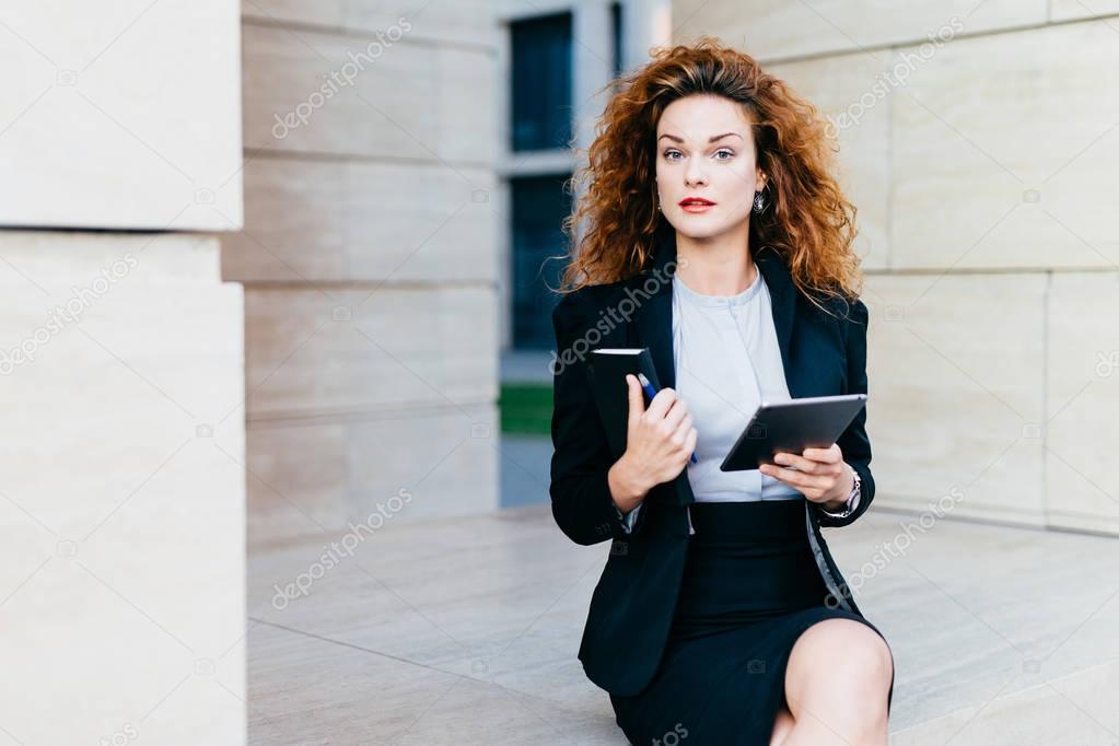 Horizontal portrait of pretty confident businesswoman dressed formally, holding her pocket book with pen and tablet computer, being busy with her work. People, lifestyle, career, business concept