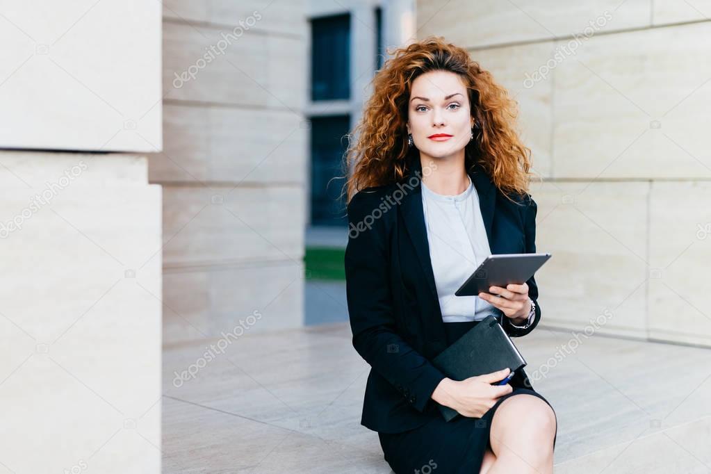 Portrait of elegant lady dressed in white blouse, black jacket and skirt, holding tablet and pocket book while sitting near office builduing. Successful businesswoman with curly hair, beautiful face