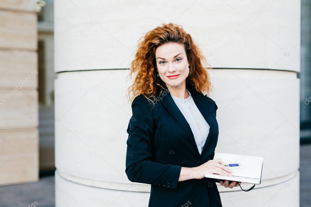 Female freelancer with bushy hairstyle, wearing white blouse, black jacket and skirt, holding her diary book, writing notes in it. Slim young lady posing against builduing. Beauty and youth concept