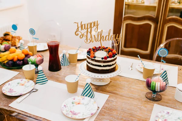 Holiday table served wit birthday cake, table full of fruit, compote, empty plates and party hats. Beautiful table set for birthday party. Festive composition. Celebration concept