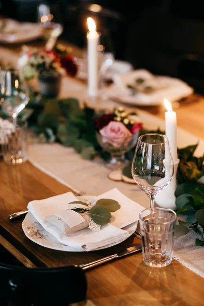 Обеденный стол накрыт для вечеринки. Тарелки, свечи, ножи, вилки на деревянном столе украшены зелеными ветвями. Стол для банкетов. Длинный праздничный стол. Концепция празднования и приема пищи — стоковое фото