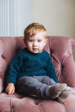 Dikey mavi eyeys ve tombul yanakları, masum küçük çocuk portresi görünüyor doğrudan kameraya, rahat pembe koltukta oturma odası, doğum günü, olduğu gibi çocuk oturur için bekleyin. Çocuk