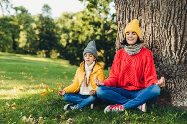 Rahat kaygısız kadın ve küçük kızı poz Lotus ağaç parkı yakınında oturmak, gözlerini kapat, konsantre, pratik yoga birlikte, sıcak giysiler giymiş çalışın. İki erkek düşünmek hakkında parça açık