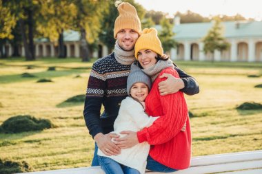 Outdoor portrait of beautiful smiling woman, handsome man and their little cute daughter stand together against ancient bilduing in park, wear warn knitted clothes, have interesting excursion clipart