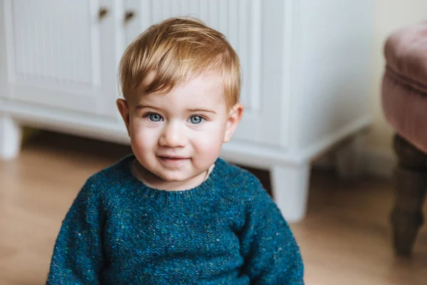 Close up retrato de criança loira com bochechas gordas, olha com olhos azuis na câmera, tem aparência inocente. Bebê adorável tem expressão séria senta-se em casa interior. Conceito de infância — Fotografia de Stock