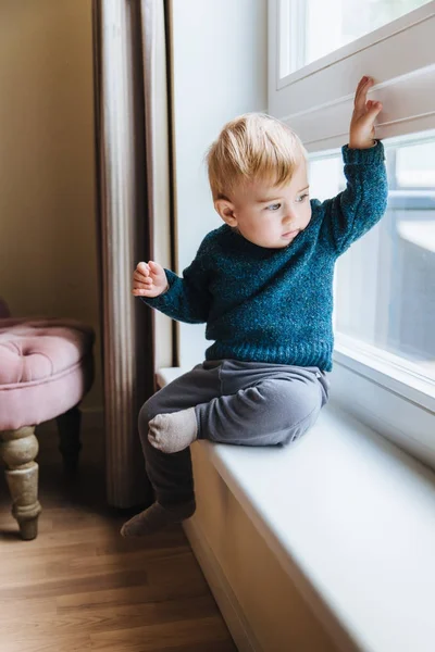Naughty small kid with blonde hair and blue eyes, sits on window sill, looks out of window, notices something in yard. Curious cute child plays alone, looks outside with attentive expression — Stock Photo, Image