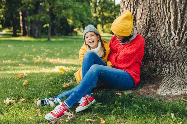 Criança engraçada e sua mãe se sentam juntos perto da árvore, caminham no parque de outono, desfrutam de momentos maravilhosos e inesquecíveis juntos. Mãe amigável e menina pequena, relaxe ao ar livre. Conceito de relaxamento — Fotografia de Stock
