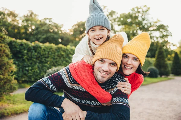 Retrato ao ar livre de homem bonito dar piggyback para sua esposa e filha, usar roupas quentes, ter expressões felizes, apoiar uns aos outros. Família abraçar ao ar livre, sorrir agradavelmente na câmera — Fotografia de Stock