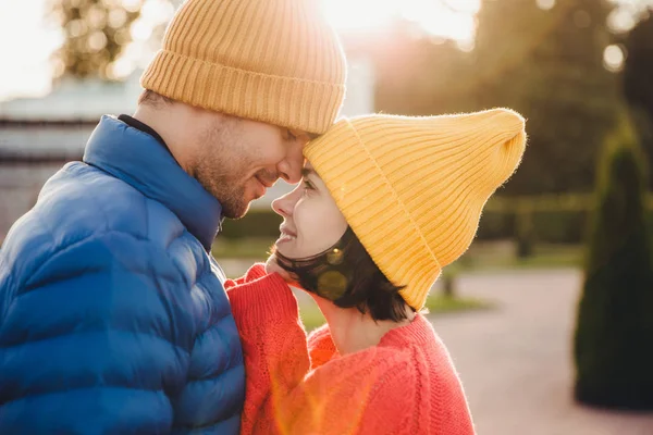 Romantic young couple look at each other with great love, have nice relationship, going to kiss, have walk outdoor in park, wear warm clothes. Lovely couple with eyes full of love and smile.