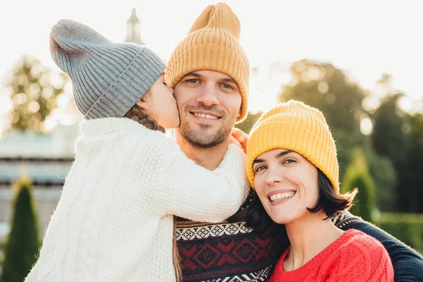 Emoções sinceras. Pequena menina bonito em chapéu de malha e suéter quente branco beija seu pai com amor. Amigável casal afetuoso posar juntos ao ar livre, sorrir feliz, ter relacionamentos maravilhosos — Fotografia de Stock