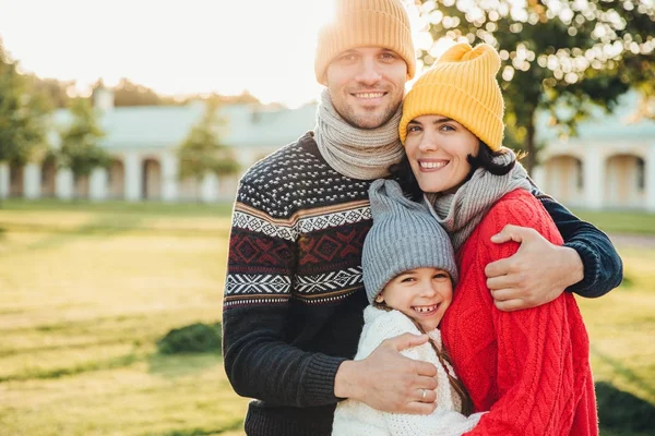 Retrato horizontal de homem bonito jovem em chapéu amarelo e camisola de malha quente, abraçar sua esposa e filha, posar na câmera enquanto ficar no parque. Três membros da família passam os fins de semana juntos — Fotografia de Stock