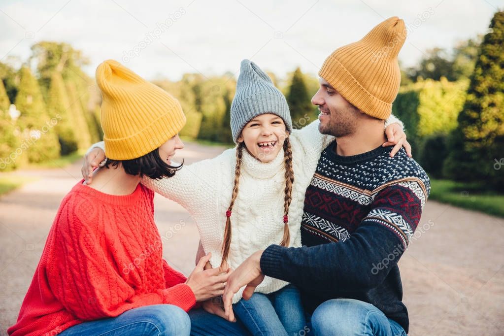 Happy small girl embrace her mother and father, have fun together, walk in beautiful park or on road. Parents look at her lovely daughter, try to do best for her. Happy family have good rest outdoors