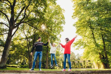 Dost sevgi dolu aile portresi birlikte eğlenin, boş zamanlarını açık havada, park yeşil ağaçların karşı durmak, rock küçük çocuk elinde çalışın. Üç aile memebers doğa üzerinde oynamak