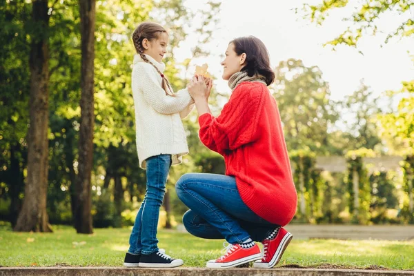 Młoda matka w ciepły sweter sweter czerwony odgrywa z jej małą córeczką w parku, daje jej liść, cieszyć się słoneczna pogoda jesienią. Affectionate Mama i dziecko spędzić razem czas odkryty — Zdjęcie stockowe