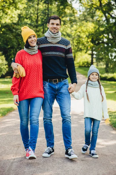 Retrato de passeio familiar amigável através do parque. Mulher bonita morena em chapéu amarelo de malha e suéter, homem bonito abraça sua esposa e segura a mão da filha, desfrutar de clima de outono e ar fresco — Fotografia de Stock