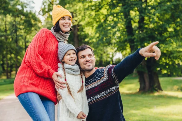 Três membros da família passam o tempo juntos, olham para o belo lago no parque, indicam com dedos, estando de bom humor, sorriem agradavelmente. Pai, mãe e filha desfrutam de união, atmosfera calma — Fotografia de Stock