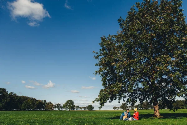 Portrait of beautiful nature and family who has picnic in background sits under big tree. Blue sky and green grass. Charming autumn weather and landscapes. Three people drink tea outdoors on meadow