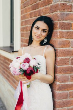 Çekici kadın gelin beyaz gelinlik giyer, buket tutar, tuğla duvara yakın duruyor, çekici bir görünüm vardır. Makyaj, zarif genç kadın vardır çekici gülümseme ona düğün kutluyor