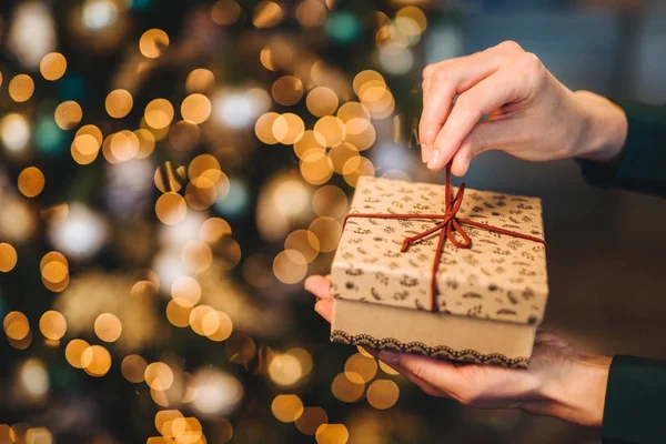 Mujer irreconocible envuelve caja de regalo sobre el árbol de Navidad con luces brillantes y guirnaldas. Caja de regalo decorada en manos de mujer. Vacaciones y concepto de celebración . —  Fotos de Stock